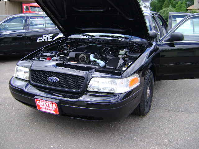 Black 2003 Ford Crown Victoria, hood open showing engine. Another black car with 'CRE' visible is in the background