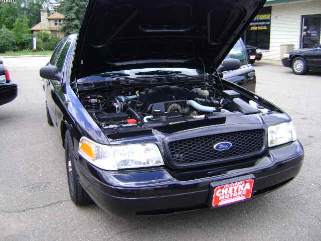 Black 2003 Ford Crown Victoria front view, hood open, showing engine. License plate frame reads CHEYKA MOTORS