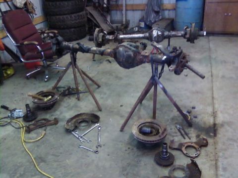 1995 Ford Bronco - Two vehicle axles are displayed on stands in a garage setting, surrounded by various tools and disassembled parts.