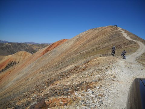 1986 Ford Bronco II - Motorcycles navigate a steep mountain road, with a vehicle visible at the summit. The landscape is arid and rocky.