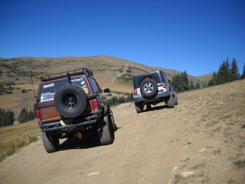 1986 Ford Bronco II - Two off-road vehicles ascend a steep, dusty mountain trail under a clear blue sky.