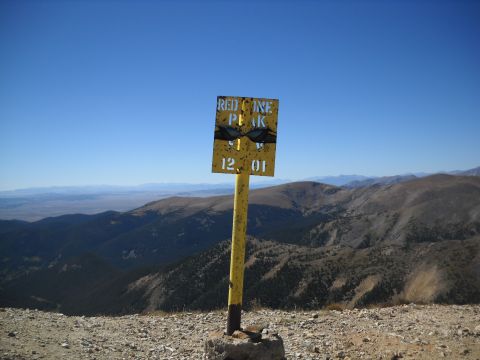 1986 Ford Bronco II - A sign marks the summit of Red Cone Peak, indicating an elevation of 12,010 feet, with a vast mountain range in the background.