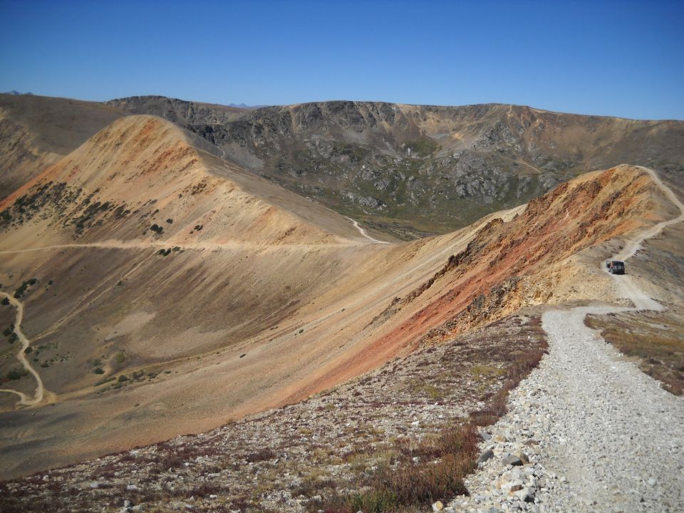 Vehicle on a dirt road winding through a mountainous landscape under a clear blue sky
