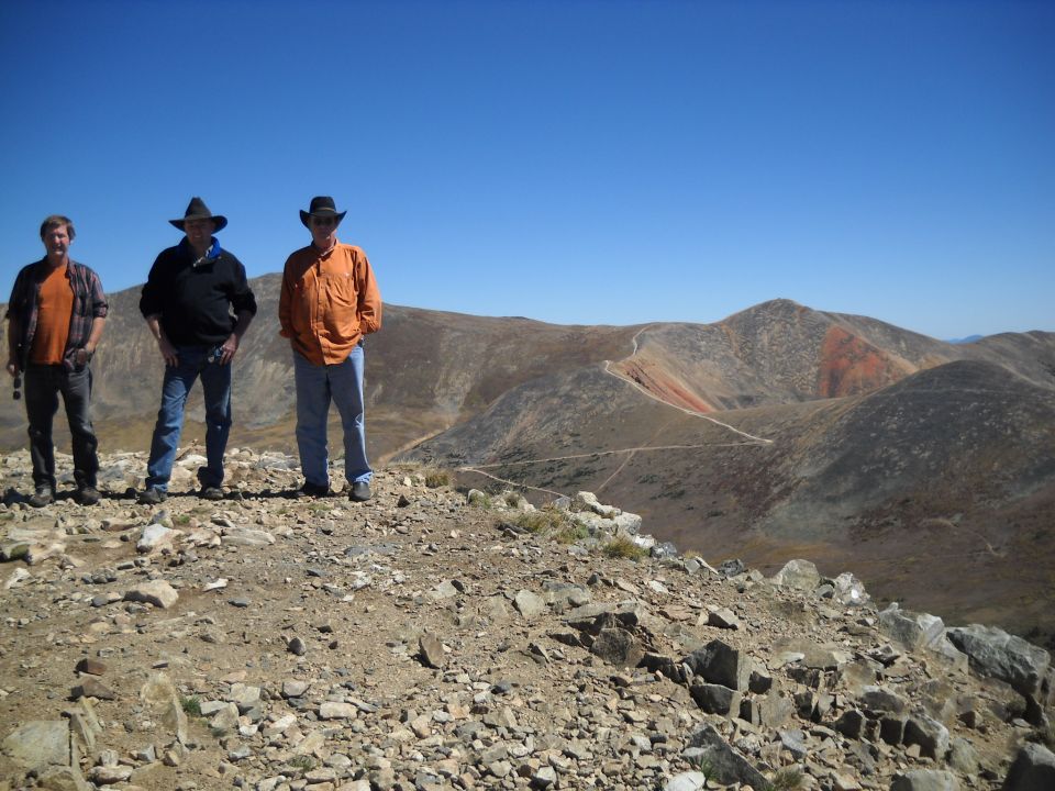 Three men in cowboy hats stand on a rocky mountain peak overlooking a vast, arid landscape with winding roads