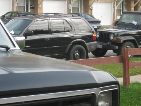 1981 Ford Bronco - A black SUV and a black Jeep are parked in a driveway in front of residential garages.