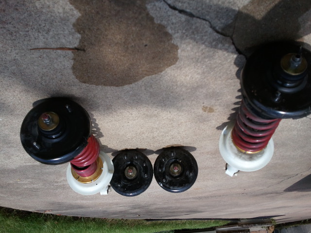 Four car suspension spring assemblies laid out on concrete. Two are complete with springs and top mounts, two are just top mounts