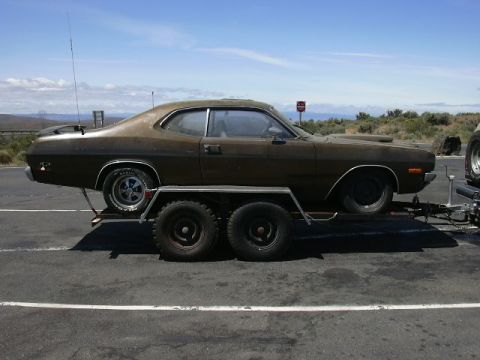 1991 Ford Bronco - Side view of a faded brown 1970s Plymouth Duster-style two-door coupe strapped to a tandem-axle open car trailer in a paved lot. The car we…
