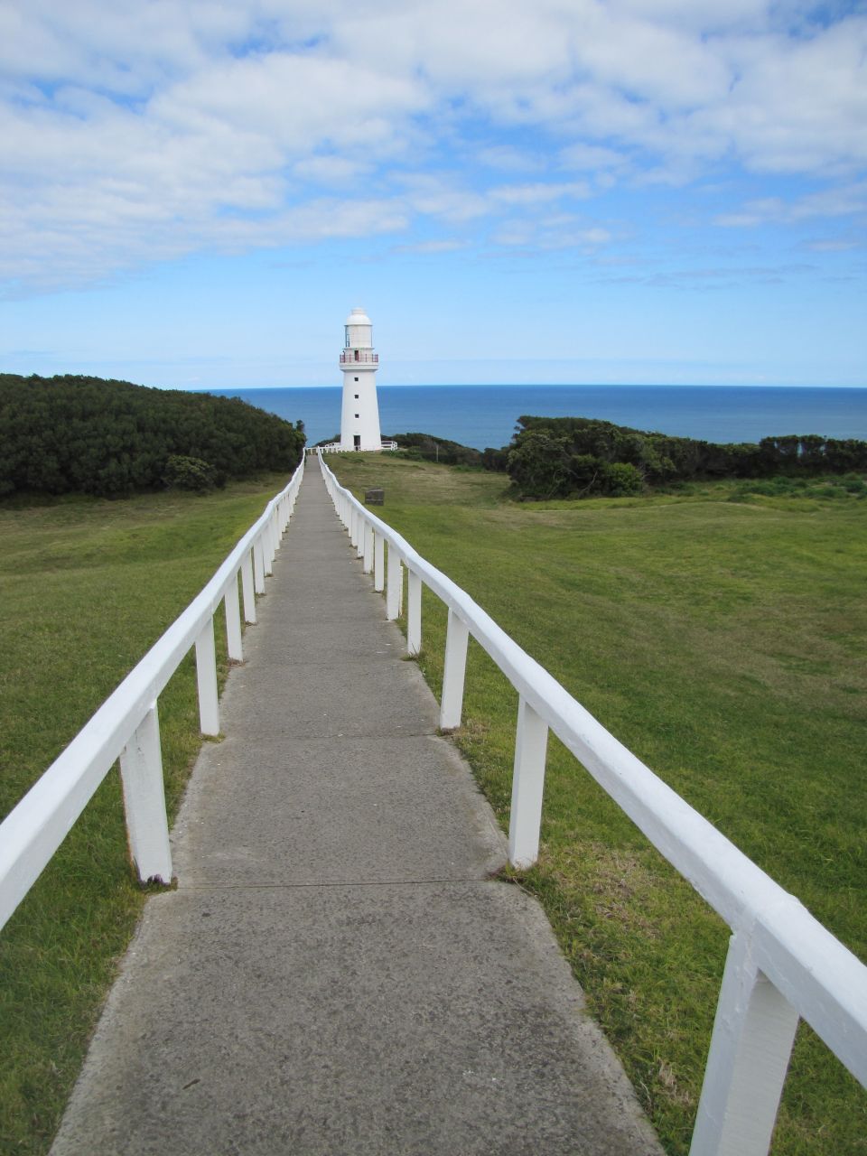White lighthouse on a grassy hill overlooking the ocean, with a paved walkway leading to it