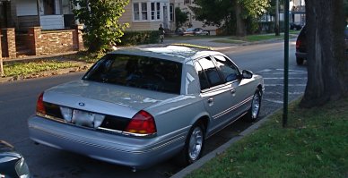 2000 Ford Crown Victoria - A silver sedan is parked curbside on a tree-lined street in front of houses.