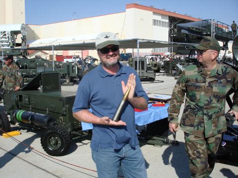 2000 Ford 427 - A man displays a large caliber bullet in front of various military vehicles and equipment.