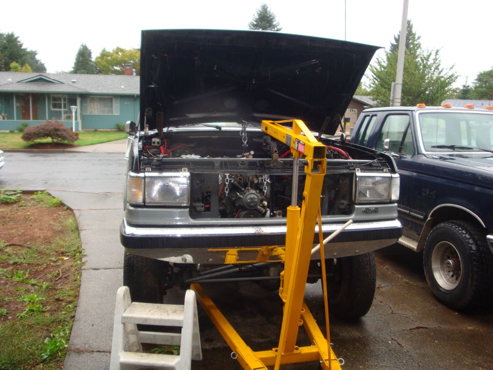 Silver and blue lifted Ford Bronco with hood up and yellow engine hoist over the engine bay