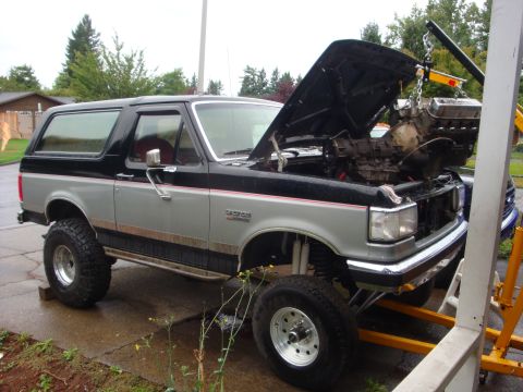 1989 Ford Bronco - A two-tone silver and black Ford Bronco has its hood open as an engine hoist prepares to remove the engine.