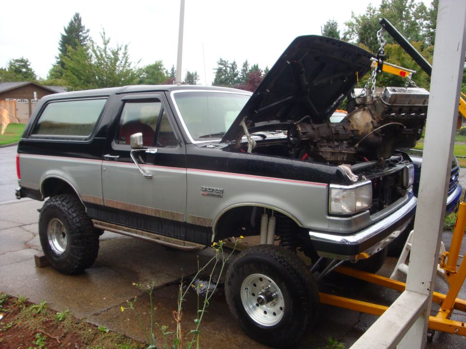Silver and black lifted Ford Bronco with engine hoisted on a crane over the open engine bay