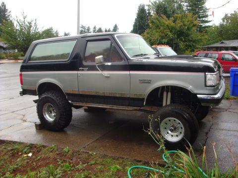 1989 Ford Bronco - A modified Ford Bronco with a two-tone paint job and raised suspension sits on a wet driveway.