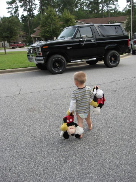 1986 Ford Bronco - A black 1986 Ford Bronco is parked on the side of a residential street, with a young child walking away from it.