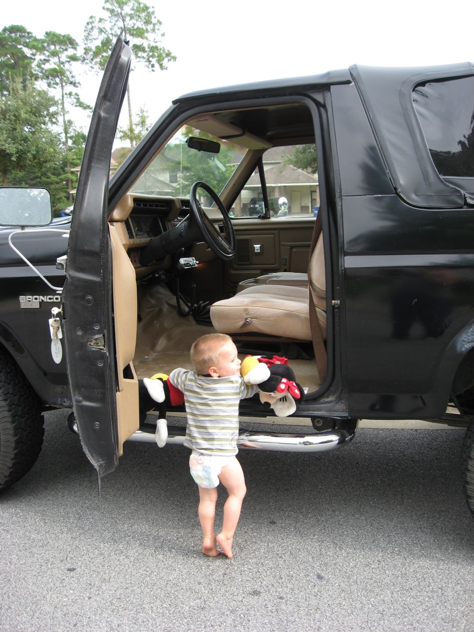 Baby in a diaper holding stuffed toys, standing by the open driver's door of a 1986 Ford Bronco