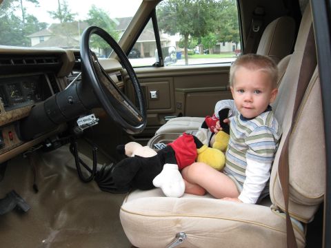1986 Ford Bronco - A young child sits in the passenger seat of a 1986 Ford Bronco, surrounded by stuffed toys.