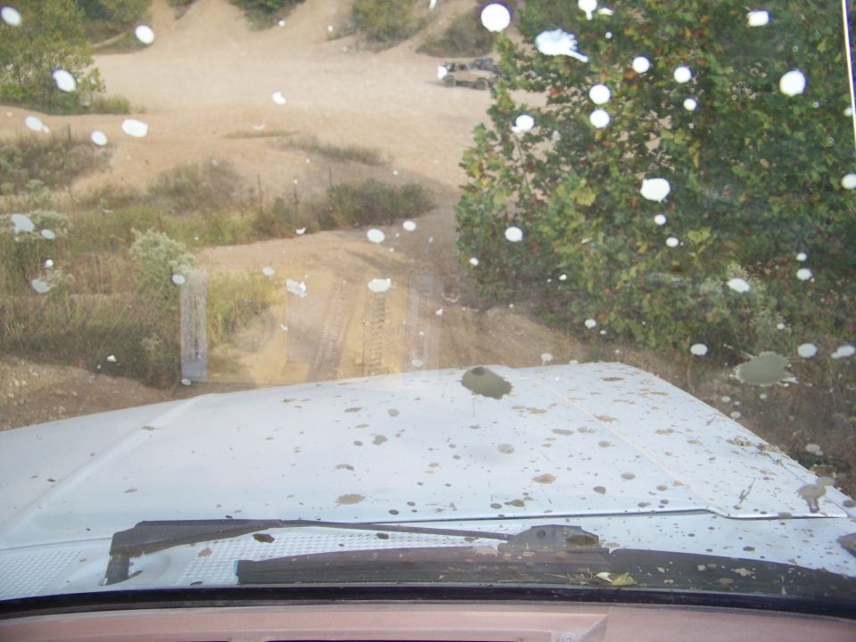 View from inside a white vehicle with a muddy windshield looking out at a dirt road and trees. Another vehicle is visible in the distance