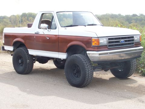 1989 Ford Bronco - This lifted Ford Bronco is parked on a dirt road with trees in the background.