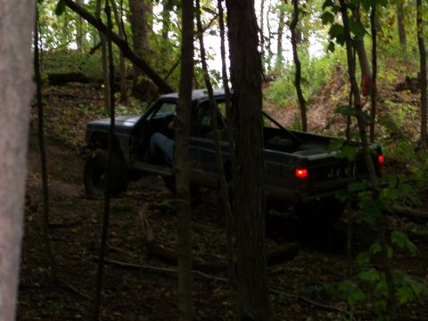 1989 Ford Bronco - A Jeep Comanche is driven through a dense forest on an uneven dirt path.