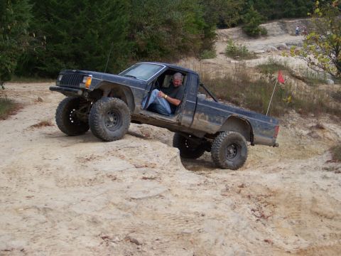 1989 Ford Bronco - Pickup truck with lifted suspension and off-road tires driving on a dirt track.
