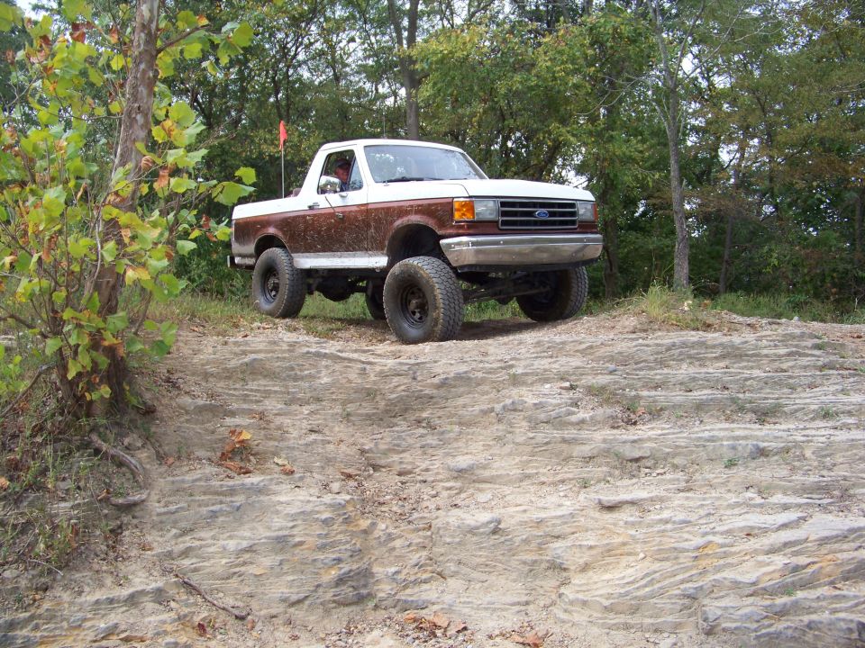 1989 Ford Bronco with brown and white paint, lifted suspension, and off-road tires driving on a rocky trail