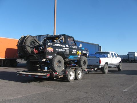 1990 Ford Bronco - black number 300 West Coast Broncos race truck strapped onto a trailer being towed by a white dually pickup in a lot.