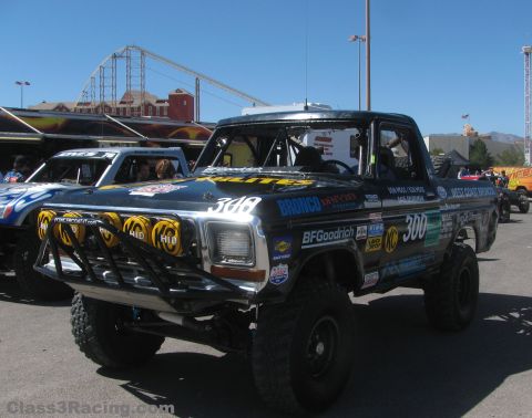 1990 Ford Bronco - black number 300 off-road Ford race truck with a light bar parked in a pit area, a roller coaster visible behind.