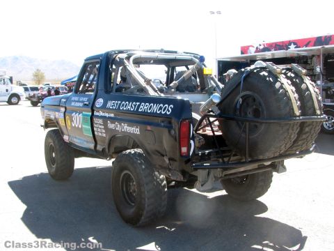 1990 Ford Bronco - Rear three-quarter view of a black number 300 West Coast Broncos race truck carrying two spare tires in a desert pit.
