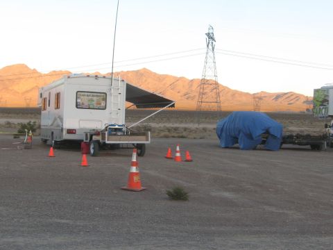 1990 Ford Bronco - RV with an awning and a tarp-covered vehicle set up in a desert pit area marked by orange cones, mountains behind.