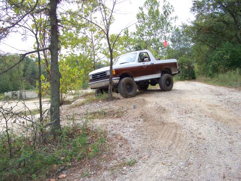1989 Ford Bronco - This lifted Ford Bronco navigates a rough, unpaved path surrounded by trees.