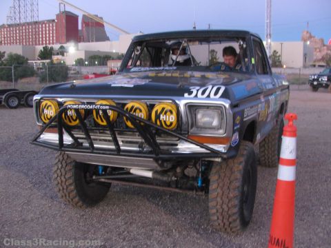 1990 Ford Bronco - Front three-quarter view of a black number 300 off-road Ford race truck with yellow HID lights, driver seated inside.
