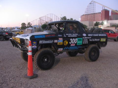 1990 Ford Bronco - black number 300 West Coast Broncos race truck staged at dusk in a gravel pit with a roller coaster in the background.