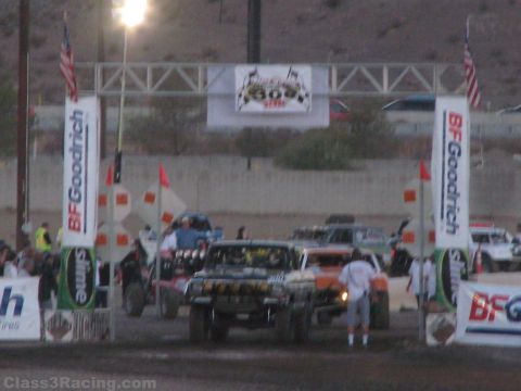 1990 Ford Bronco - Off-road race trucks line up under a banner-topped start arch flanked by BFGoodrich flags and American flags at a desert track at dusk.