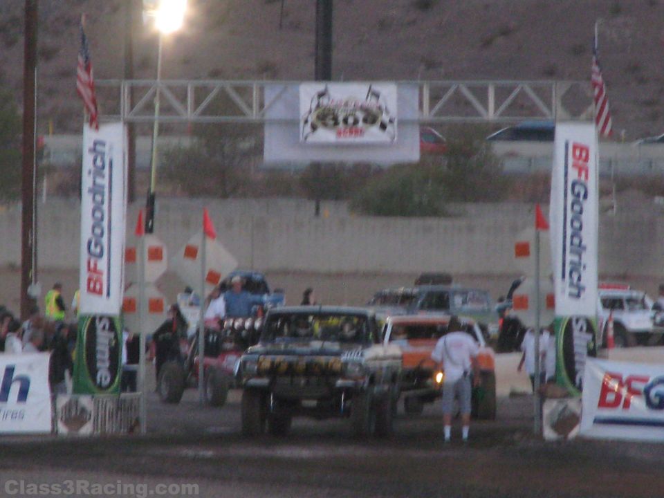 Off-road race trucks staged under a lit BFGoodrich start arch at dusk