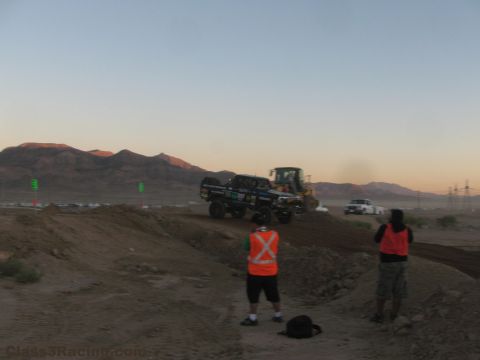 1990 Ford Bronco - off-road race truck crests a dirt berm beside a wheel loader as two vest-clad marshals watch against mountains at dusk.
