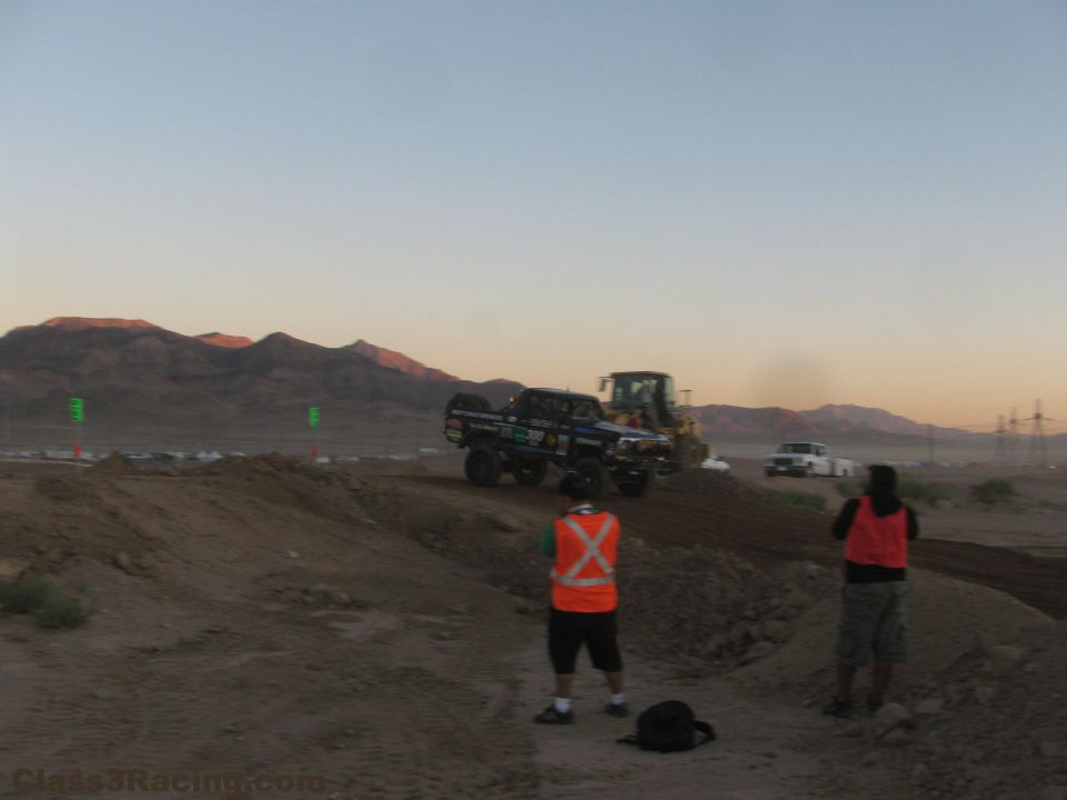 Off-road truck cresting a berm near a loader with spectators at dusk