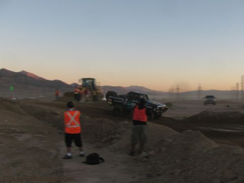 1990 Ford Bronco - number 300 off-road race truck moves along a dirt course at dusk as marshals in safety vests look on near a loader.