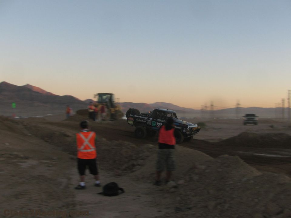 Off-road race truck on dirt course at dusk with marshals watching