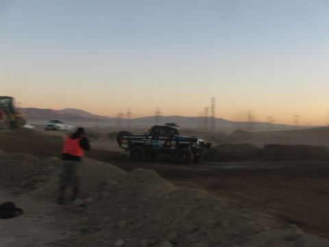 1990 Ford Bronco - black number 300 off-road race truck speeds across a desert dirt track at dusk with power-line towers on the horizon.