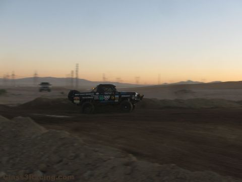 1990 Ford Bronco - black number 300 off-road race truck launches over a dirt berm at dusk with power lines and hills in the background.