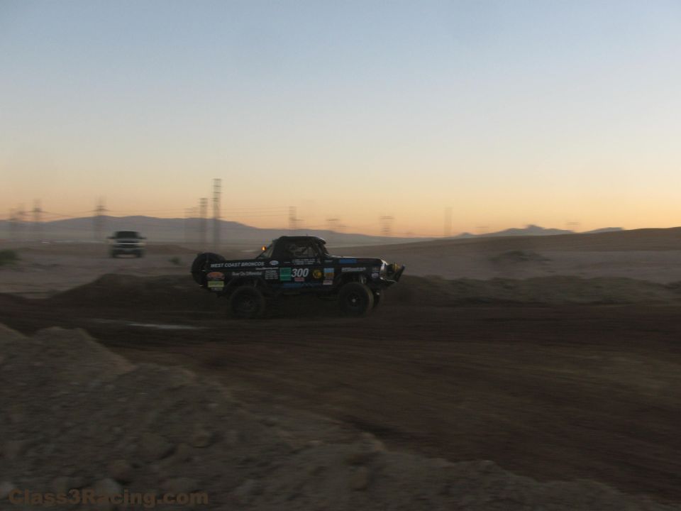 Number 300 off-road race truck airborne over a berm at dusk