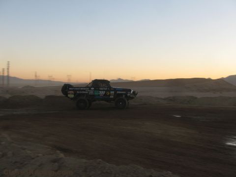 1990 Ford Bronco - West Coast Broncos number 300 off-road race truck drives across a desert dirt track at dusk near transmission towers.