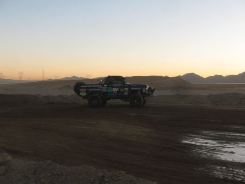 1990 Ford Bronco - black number 300 West Coast Broncos race truck travels a desert dirt course with mountains and a fading sunset behind.