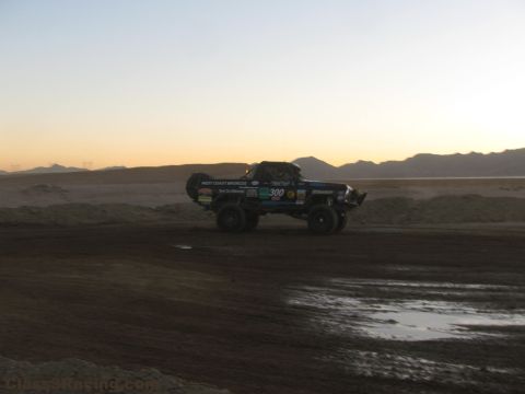 1990 Ford Bronco - number 300 off-road race truck moves along a desert dirt track at sunset with mountain ranges in the background.