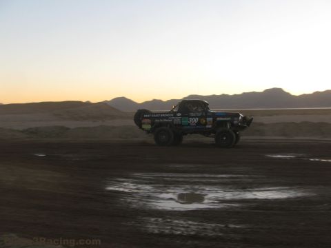1990 Ford Bronco - West Coast Broncos number 300 off-road race truck is silhouetted against a sunset over a desert dirt course.