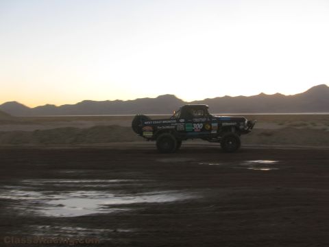 1990 Ford Bronco - number 300 off-road race truck drives across a damp desert dirt track with low mountains under a fading sky.