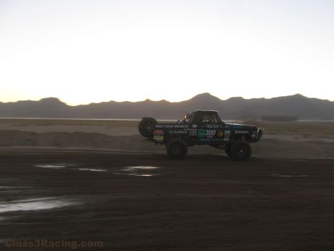1990 Ford Bronco - number 300 West Coast Broncos race truck catches air over a desert dirt course with mountains on the horizon at dusk.