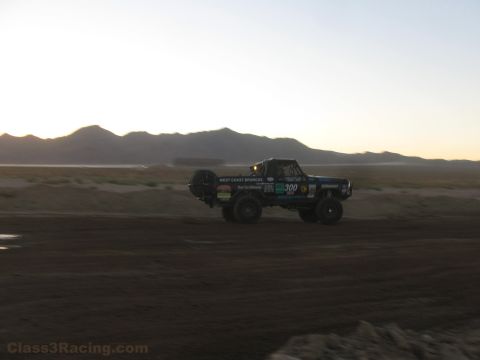 1990 Ford Bronco - number 300 off-road race truck speeds along a desert dirt course with a spare tire on the bed and mountains behind.