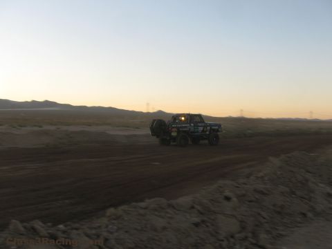 1990 Ford Bronco - off-road race truck drives away along a wide desert dirt road at dusk with hills and power lines in the distance.